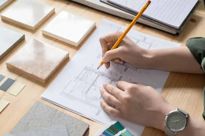Hands drawing a floor plan in pencil beside material and finish samples.